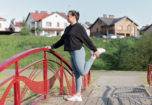 A woman in active wear performs a stretching exercise on a red bridge in a suburban park setting.